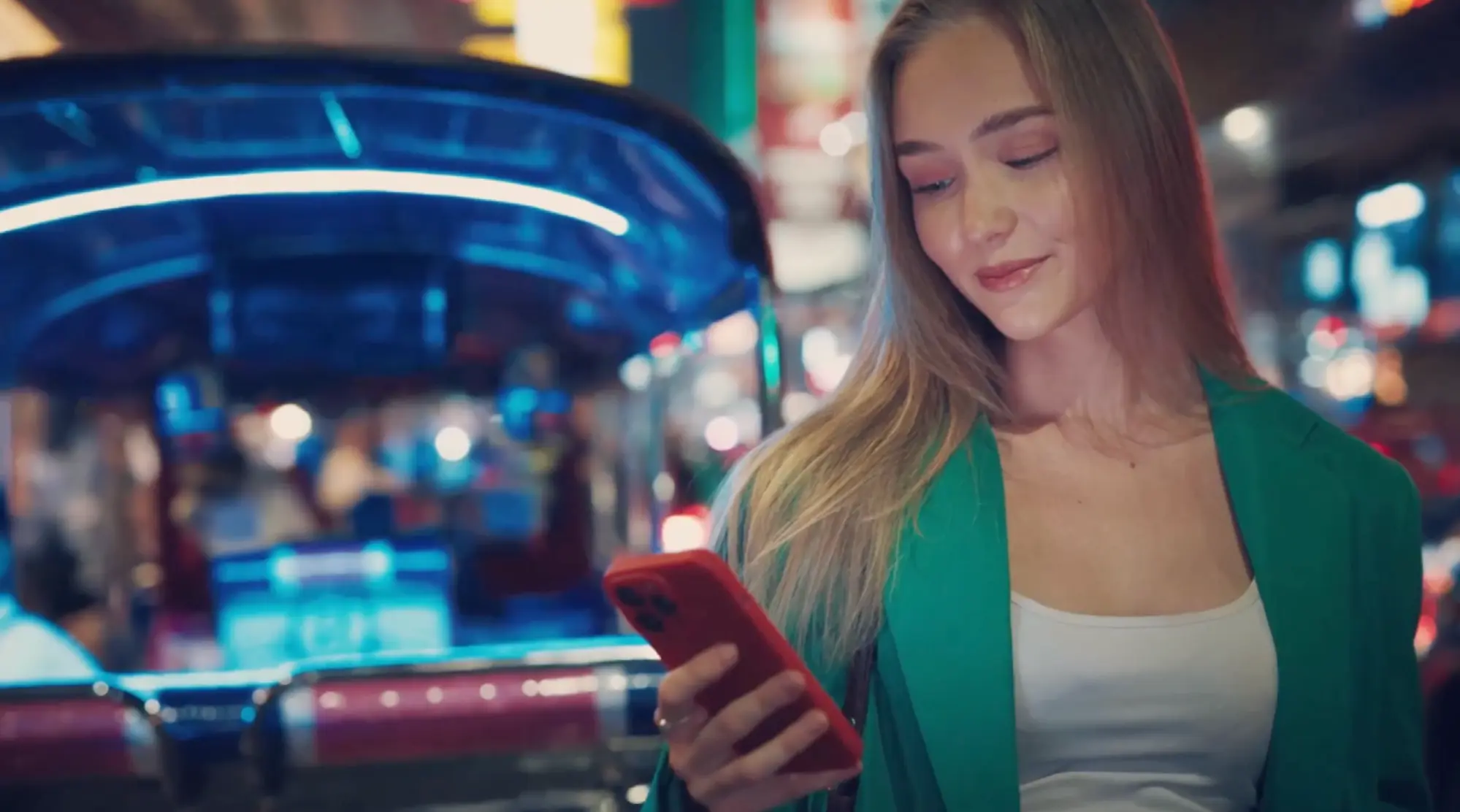 Mladá žena v zeleném saku se dívá na červený telefon na noční ulici s neonovými světly.